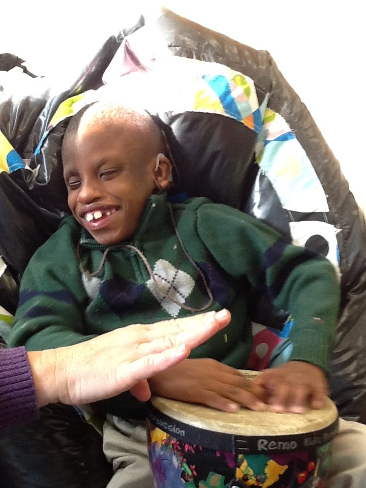 Young boy sitting in a wheelchair, holding a tambourine on his lap and touching it with both hands. Big smile on his face.